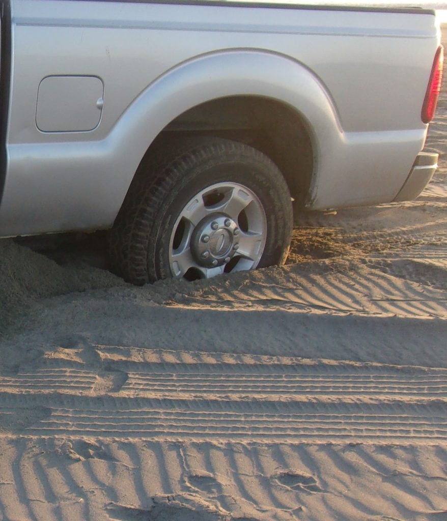 A truck, badly stuck in the sand on a beach.